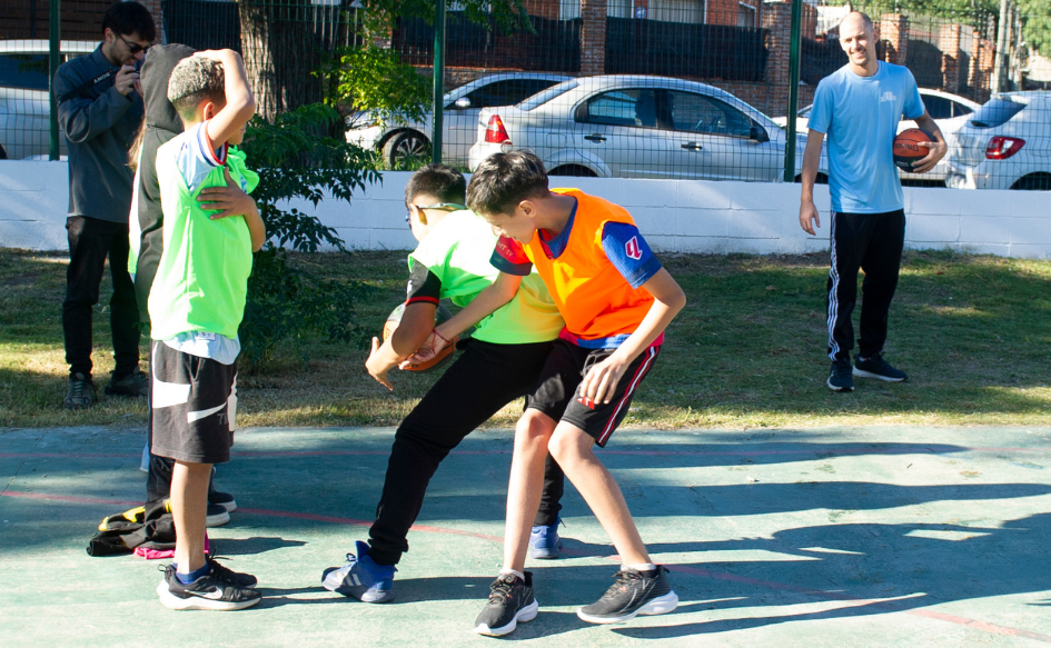 Niños de unos 10 años se disputan una pelota de básquetbol. Detrás, los observa un docente con una pelota en la mano