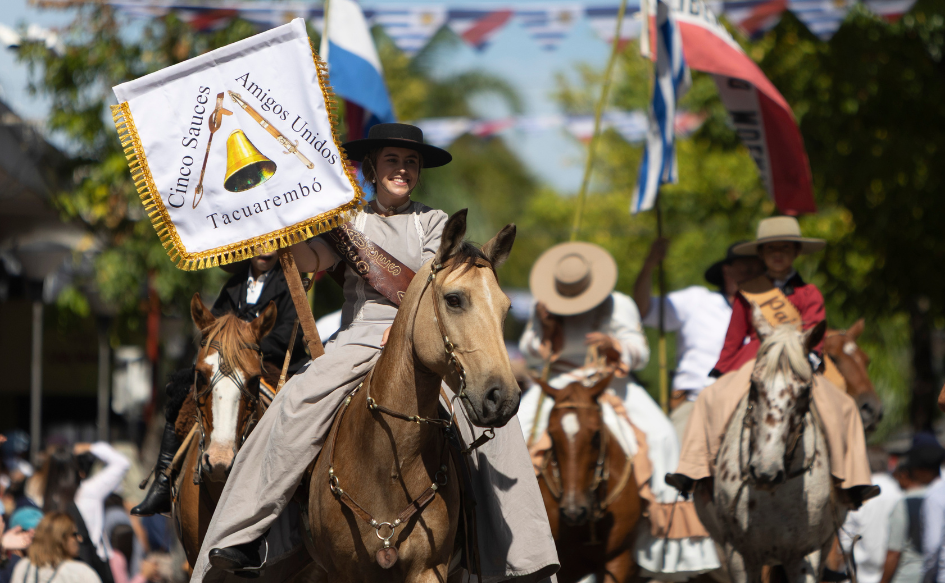 Mujer de unos 20 años, desfila montando a caballo, con un cartel con la leyenda "Cinco Sauces, Amigos Unidos". Detrás, un conjunto de jinetes con los pabellones patrios. 