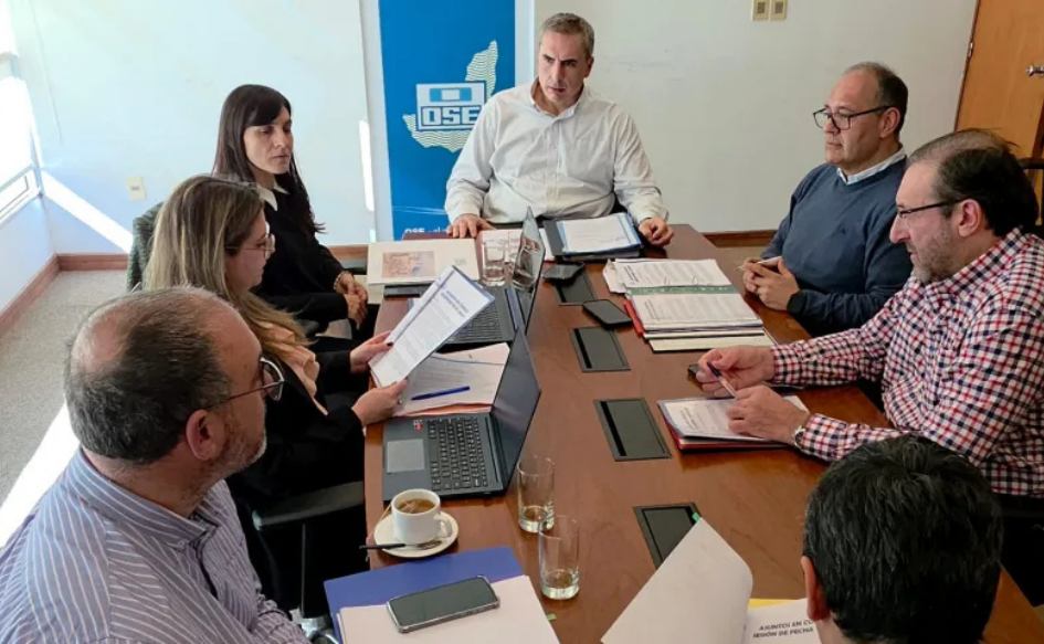 Directorio de OSE, encabezado por su presidente, Pablo Ferreri, reunido en una oficina en el departamento de Maldonado. Los integrantes están sentados alrededor de una mesa, en la que hay documentos en papel y notebooks. Detrás, un banner con el logo de OSE