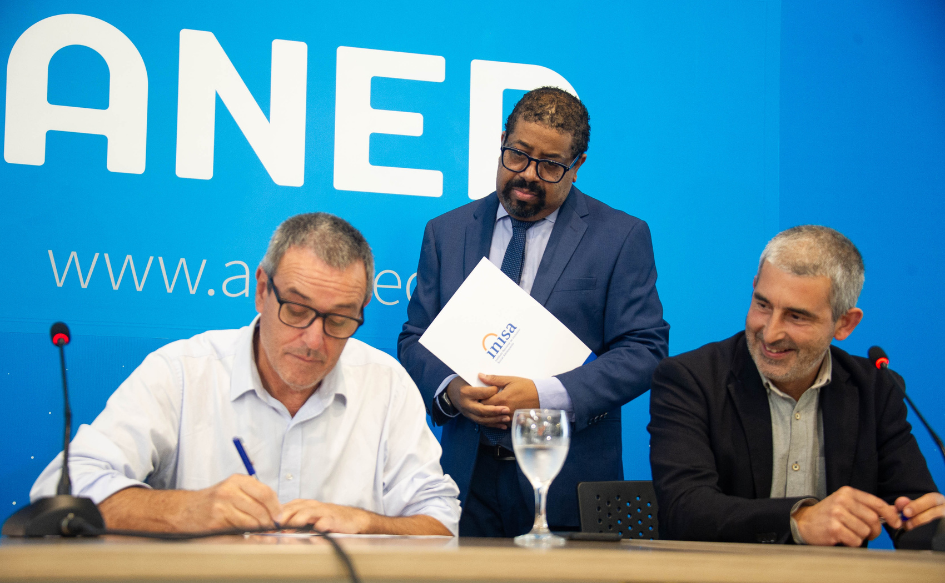 Presidente de Inisa, Jaime Saavedra, firma un documento. El presidente de ANEP, Pablo Caggiani, sentado a su lado, lo observa sonriente. Detrás, un funcionario de pie, con una carpeta con el logo de Inisa