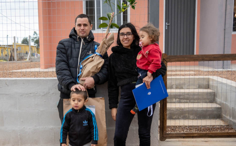 Una pareja con dos niños, miran a la cámara desde el portón de su casa, recibida a través del plan Juntos. El hombre sostiene una planta y apoya su mano en la cabeza de un niño de unos seis años. La mujer, sostiene una niña de unos tres años y una carpeta con el logro del Ministerio de Vivienda
