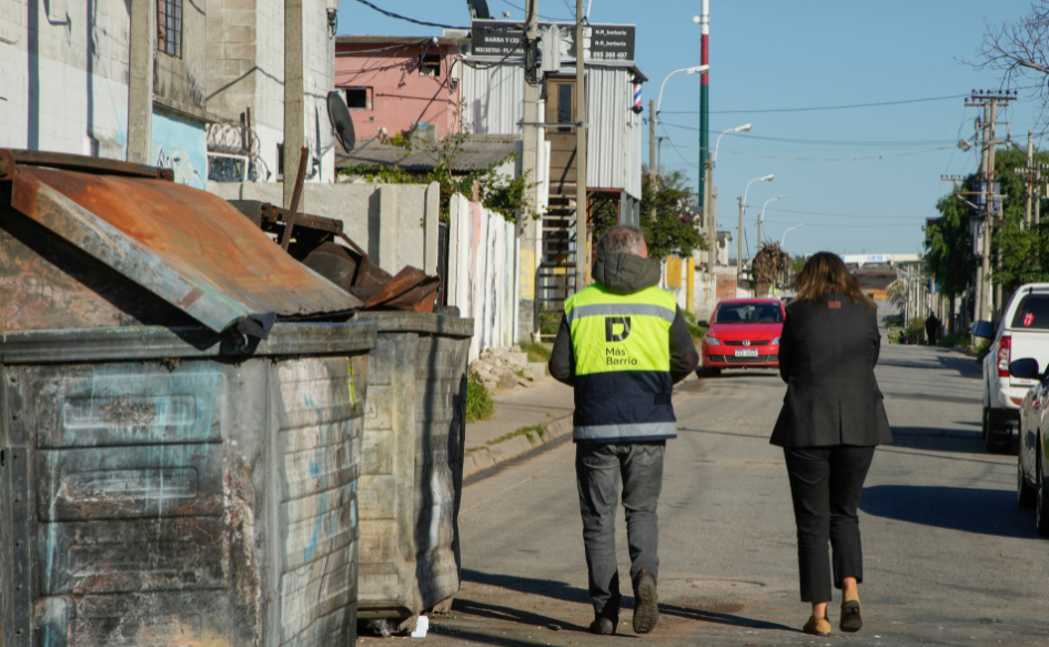 Funcionario del Mides, identificado con chaleco amarillo y el logo de Más Barrio, camina por una calle de Cerro Norte, junto a la ministra de Vivienda, Tamara Paseyro. 