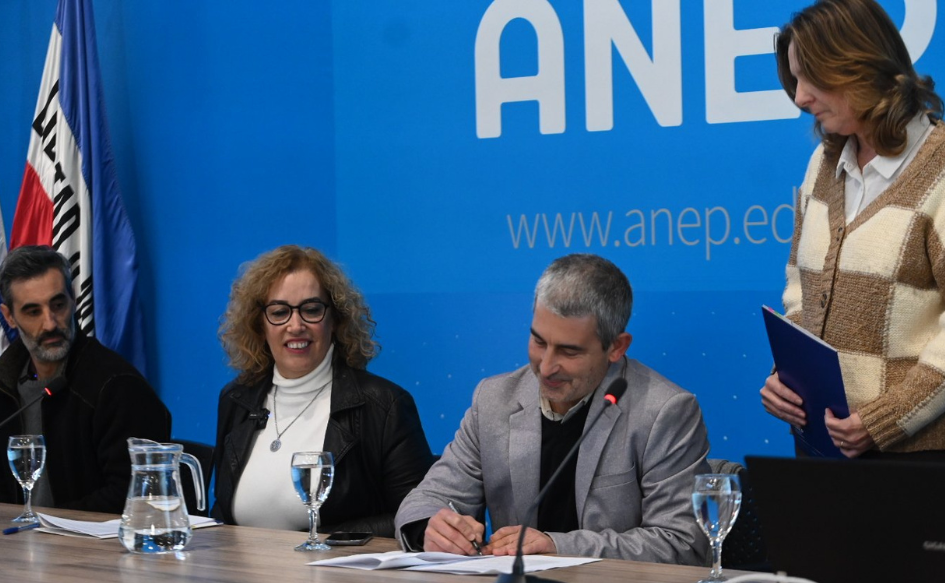 Presidente de ANEP, Pablo Caggiani, firma un documento. A su lado, lo observan autoridades de la Red de Centros. Detrás de él, una funcionaria sostiene una carpeta