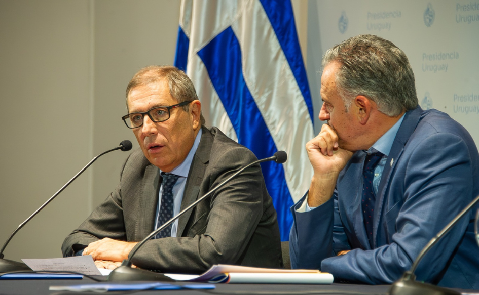 Ministro de Economía, Gabriel Oddone, de lentes, habla en conferencia de prensa. A su lado, el presidente Yamandú Orsi lo observa con su mano en el mentón. De fondo, el pabellón nacional