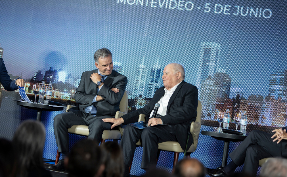 Miembro de la Fundación Euroamérica, Enrique Iglesias, sentado en un sillón, habla apoyándo su mano en la pierna del presidente Yamandú Orsi, quien lo mira sonriente, sentado a su lado, con la mano derecha apoyada sobre su antebrazo izquierdo