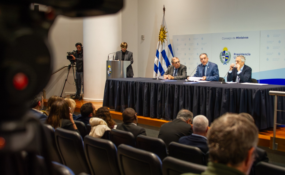 Ministros observan la conferencia de prensa desde la primera fila. En el escenario, el presidente Yamandú Orsi habla mientras sostiene un documento en la mano. A su lado, lo observan el ministro de Economía, Gabriel Oddone y el director de OPP, Rodrigo Arim. Al costado de la mesa, la maestra de ceremonias permanece detrás de un atril y un camarógrafo registra a los ministros