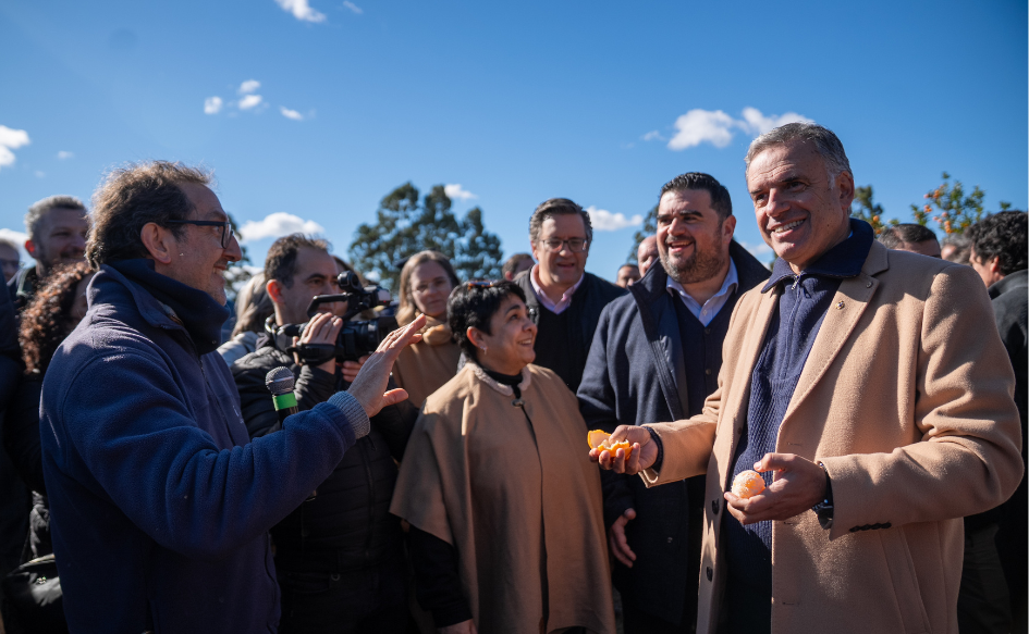 Presidente Yamandú Orsi sostiene una naranja con su mano izquierda, y la cáscara en su mano derecha. A su lado, un periodista le habla sonriente. Atrás, el intendente de Paysandú, Nicolás Olivera y una decena de personas, sonríen