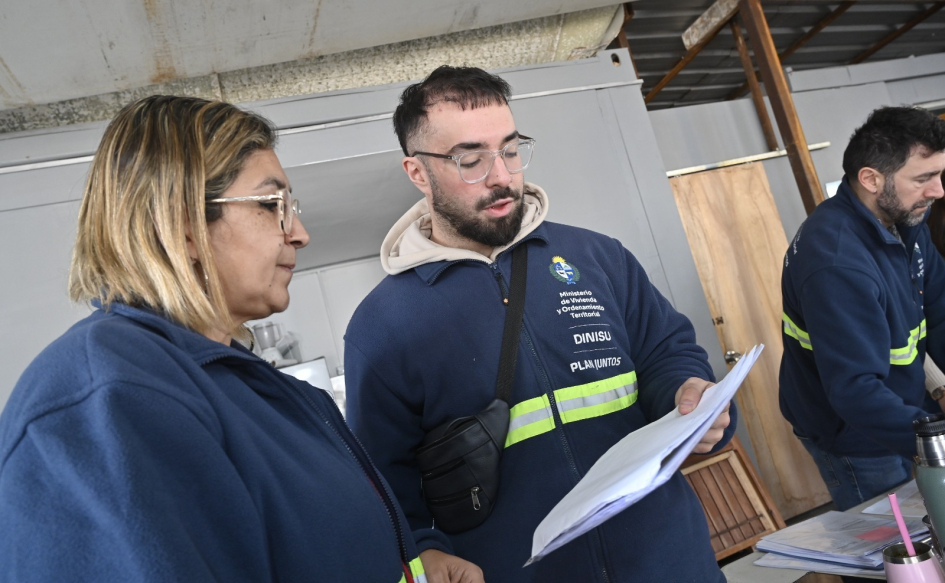 Un funcionario y una funcionaria identificados con ropa de la Dinisu del Ministerio de Vivienda, observan una hoja sostenida por uno de ellos, en una casa afectada por el temporal del 18 de julio en Salto