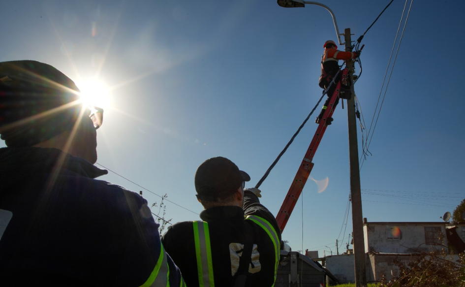 Un funcionario de la Intendencia de Montevideo manipula tendido eléctrico subido a una escalera, mientras un funcionario, desde el piso, sostiene un cable y otro lo observa