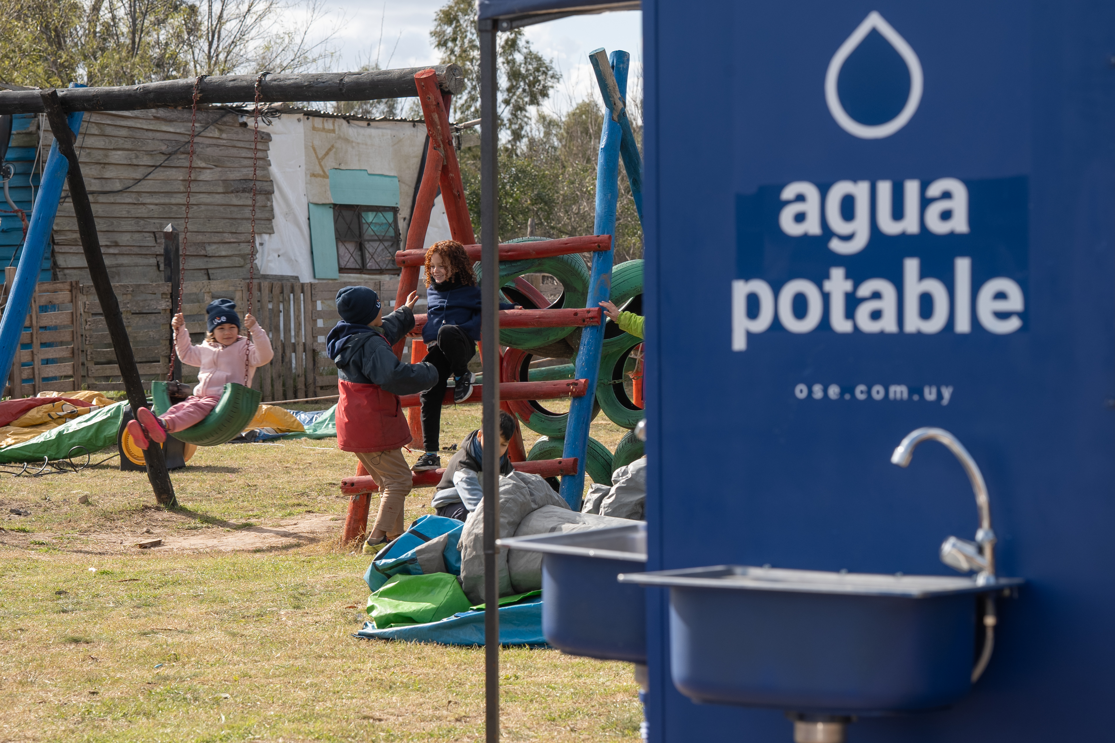 Niños juegan en hamacas de madera en el barrio Nuevo Comienzo. Delante de ellos, una pileta con una canilla y un banner que dice "agua potable"