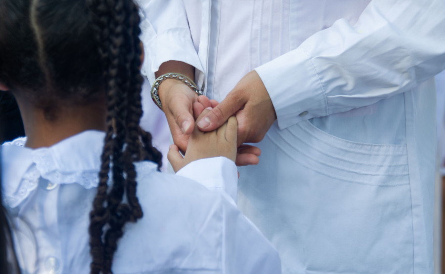 Niña de trenzas, con túnica, toma la mano de una maestra