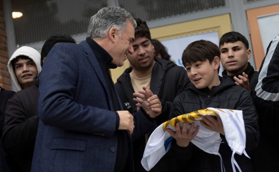 Presidente Yamandú Orsi sonríe mirando a un estudiante, que sostiene le pabellón nacional en sus manos. Detrás de él, ot