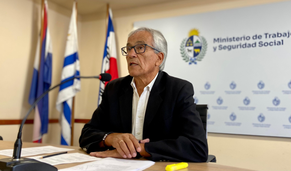 Ministro de Trabajo, Juan Castillo, habla sentado con los codos apoyados en su escritorio. En el fondo, el pabellón nacional y un banner con el logro del Ministerio de Trabajo y Seguridad Social