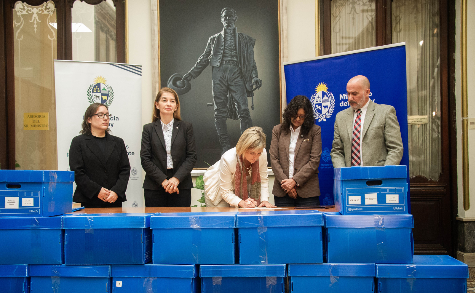 Interior entregó 73.000 páginas de microfilmación de documentos del pasado reciente a Presidencia de la República Subsecretaria del Ministerio del Interior, Gabriela Valverde, firma un documento inclinada sobre una mesa. A su lado, la observa de pie la directora de la Secretaría de Derechos Humanos para el Pasado Reciente. Enfrente, decenas de cajas.