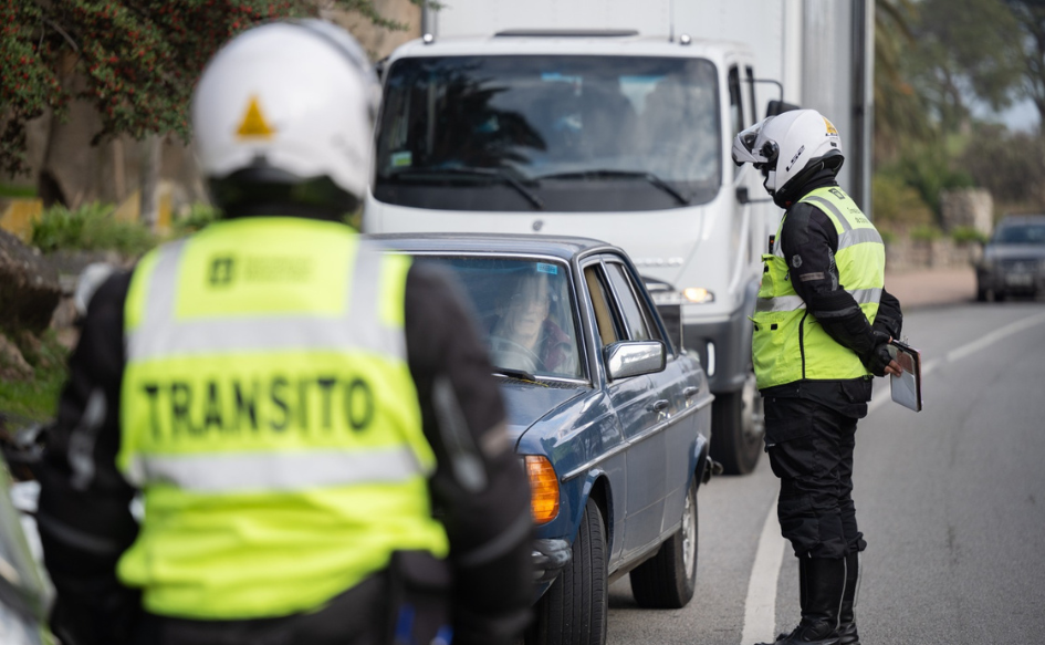 Un inspector de tránsito, de pie, dialoga con el chofer de una auto detenido en la vía pública. Delante de ellos, otro inspector los observa. En su chaleco dice "tránsito". 