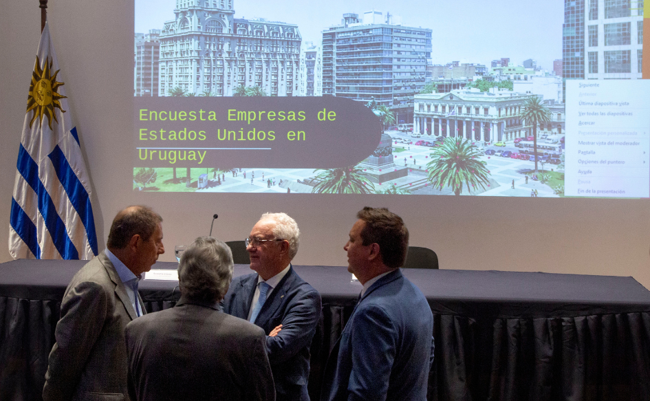 El 80% de las empresas estadounidenses destacan el buen clima de negocios en Uruguay Ministro de Economía y Finanzas, Gabriel Oddone, habla con el canciller Mario Lubetkin y dos personas más, en el salón de actos de Torre Ejecutiva. Atrás de ellos, se proyecta una imagen de la plaza Independencia con la leyenda "Encuesta empresas de Estados Unidos en Uruguay"