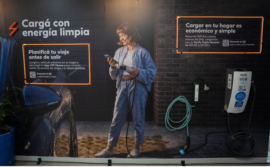 Imagen interior del stand de UTE en la Expo Prado 2026. Una mujer joven sostiene un cargador de autos eléctricos. Se lee "Cargá con energía limpia" y "Cargar en tu hogar es económico y simple"