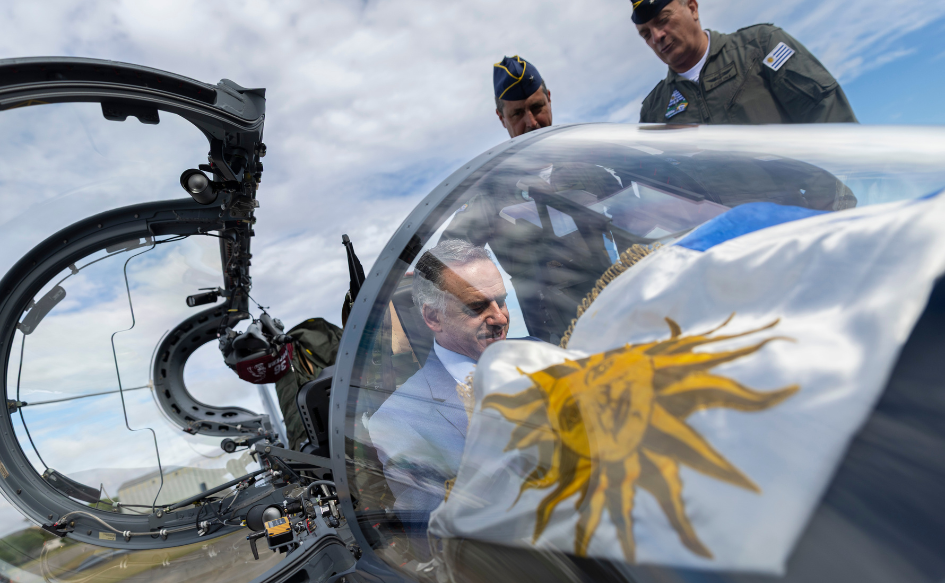 Presidente Yamandú Orsi sentado en la cabina de una aeronave Super Tucano de la Fuerza Aérea Uruguaya, con el pabellón nacional. Atrás, lo observa el comandante en Jefe de la FAU, Fernando Colina