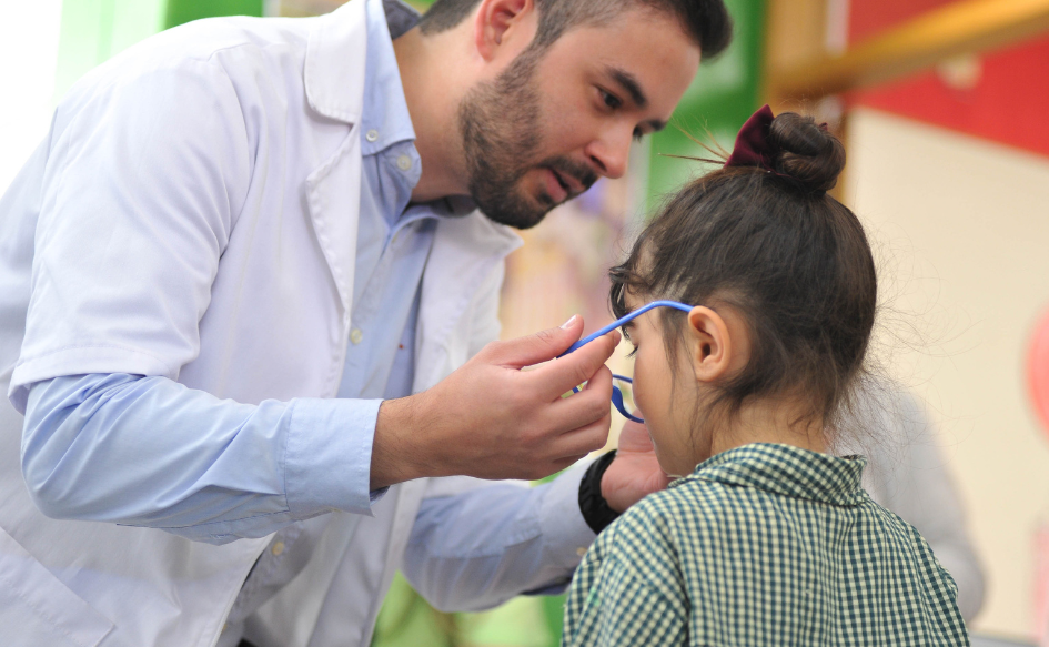 Técnico óptico, con túnica, coloca lentes a una niña de unos cuatro años, con uniforme de centro de educación inicial