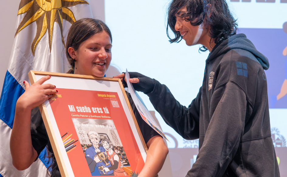Dos adolescentes sostienen un cuadro durante la premiación de los Juegos Narrativos 2026. El cuadro tiene dos personajes de historieta levantando un trofeo y la leyenda "Mi sueño eres tú"