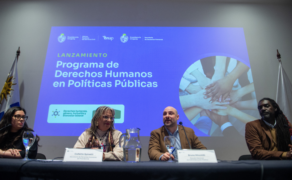 Funcionarios de la Administración Central se formarán en Derechos Humanos Director de la ENAP, Bruno Mincilli, habla en conferencia. A su lado, lo observa la secretaria de Derechos Humanos, Collette Spinetti. Atrás, se proyecta una presentación titulada "Derechos Humanos en políticas públicas" con una imagen de muchas manos, unas sobre otras.