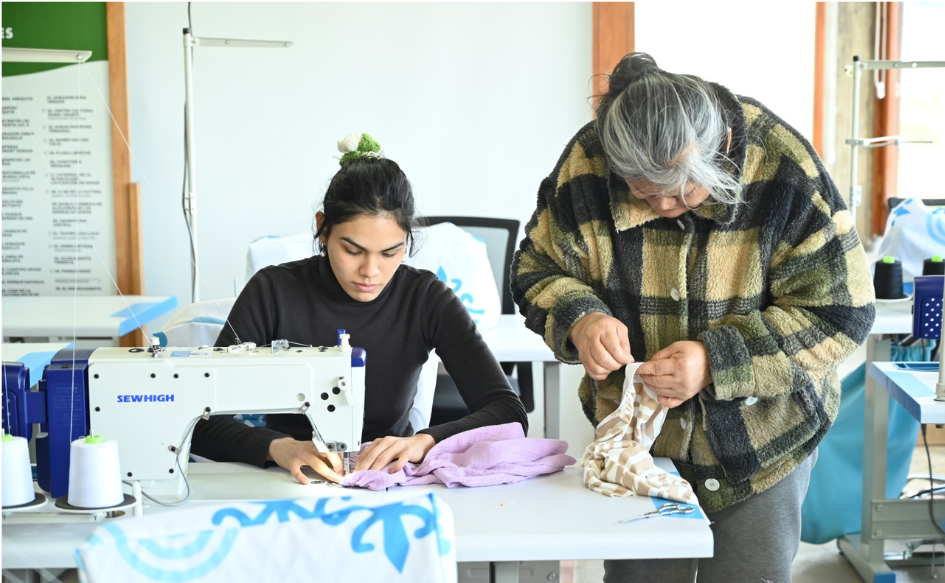 Mujer cosiendo sentada, con una máquina de coser, en el Centro de Costura Comunitario de la ciudad de Minas. A su lado, de pie, otra mujer cose a mano, con una aguja. 