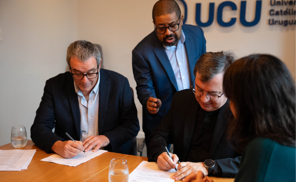 Presidente de Inisa, Jaime Saavedra, y rector de la UCU, Julio Fernández, firman el convenio marco entre ambas instituciones. Detrás de ellos, un funcionario les indica dónde tienen que firmar. En la pared, en el fondo, el logo de UCU. 