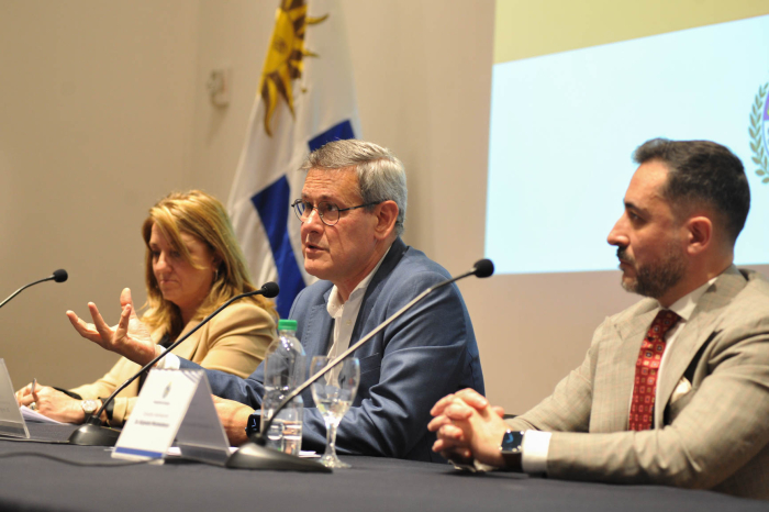 Prosecretario de Presidencia diserta sentado en mesa con micrófono en salón de actos. 