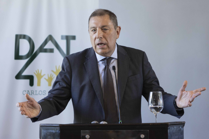 Ministro de Economía diserta parado en atril con micrófono. Atrás cartel con logo de DAT. 