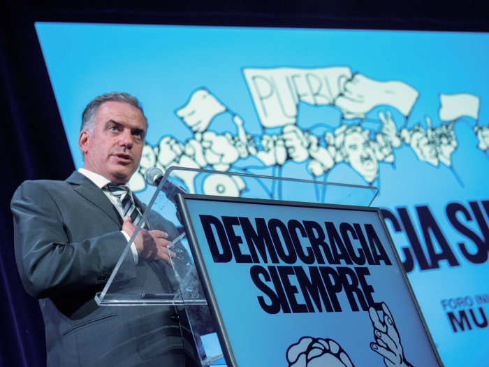 Orsi diserta, parado en atril con logo que dice democracia siempre y retrato de José Mujica