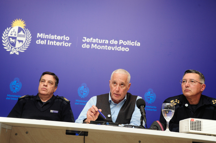 Ministro del Interior diserta sentado en mesa con micrófono junto a dos autoridades policiales. 