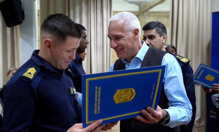 Ministro del interior le entrega un diploma azul con logo amarillo a un policía uniformado. 