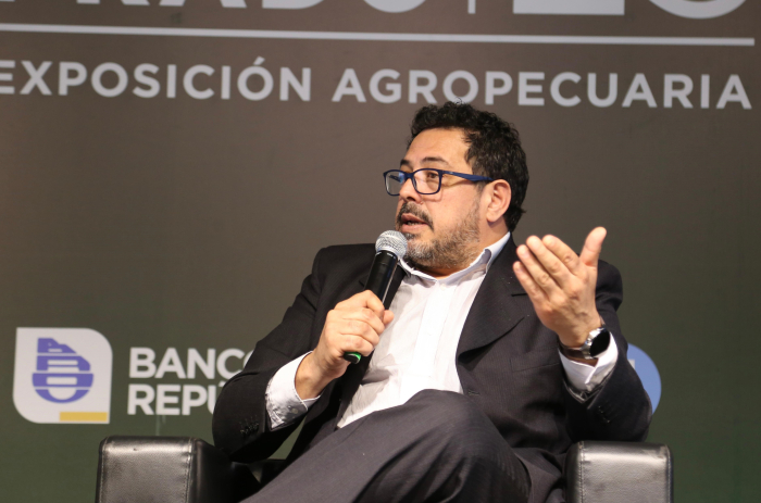 Un hombre diserta sentado en un sillón, con micrófono en mano, junto al secretario de Presidencia. 