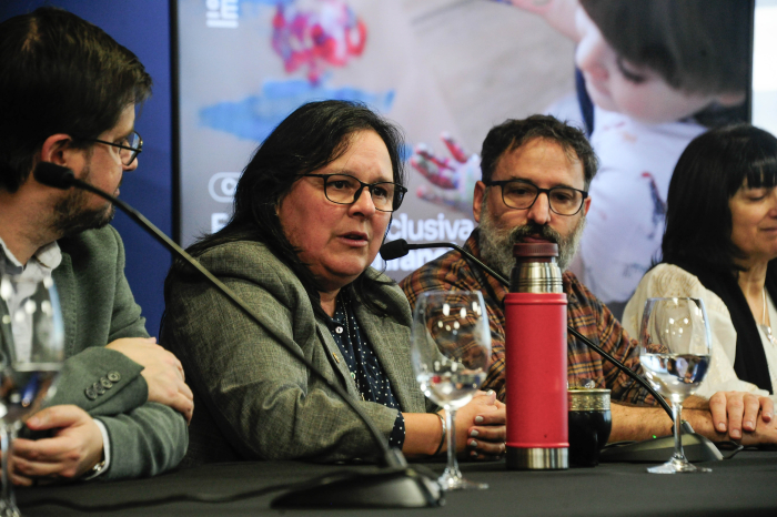 Hombre diserta, sentado en mesa con micrófono, junto a la ministra interina de Educación