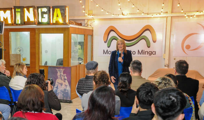 Cosse diserta de pie con micrófono en mano frente a público sentado en salón de eventos. 