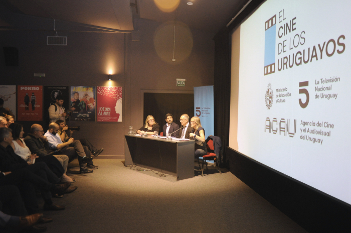 Plano general del auditorio con una gran pantalla en el centro, una mesa de cuatro oradores 