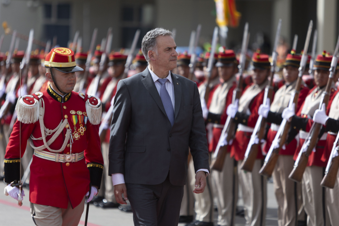Orsi vestido de traje oscuro camina con paso firme junto a un oficial militar uniformado de rojo