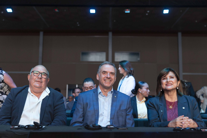 Orsi sonriente junto a dos participantes del evento sentado en mesa mira hacia adelante.