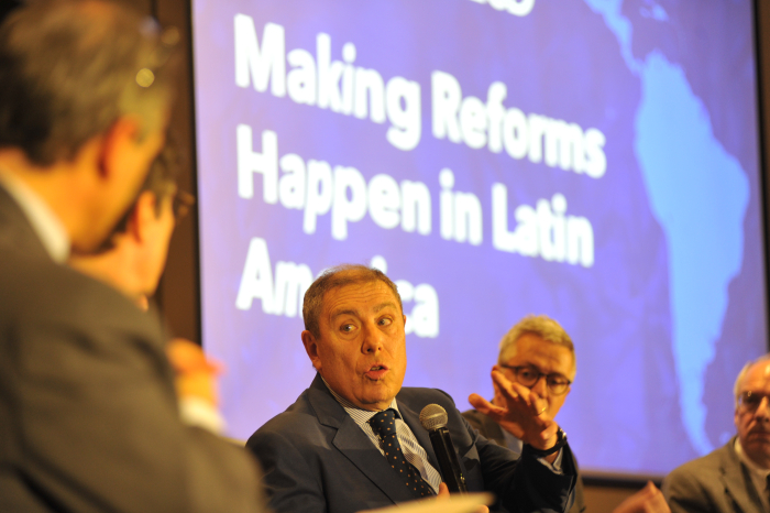 Ministro de Economía diserta sentado en mesa con micrófono en mano