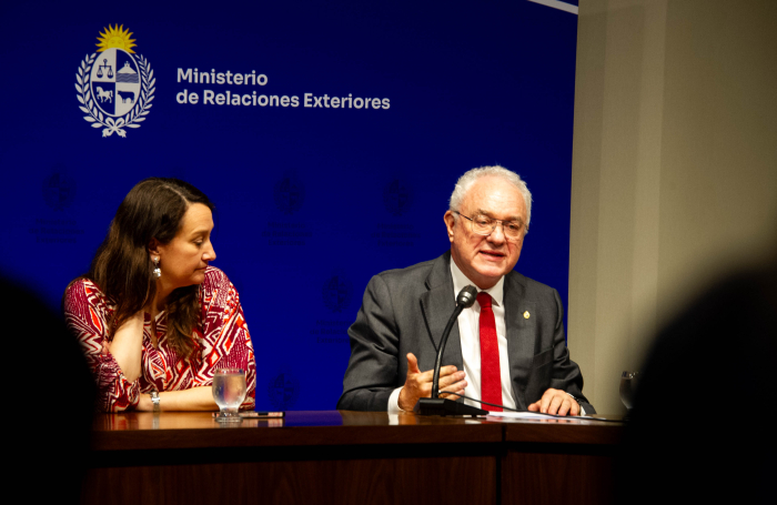 Canciller diserta sentado en mesa con micrófono, a su lado está la vicecanciller.