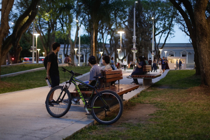 Dos jóvenes están sentados en un banco de la plaza 19 de Abril, en Treinta y Tres. Frente a ellos, un joven de pie. Sobre el banco, una bicicleta apoyada