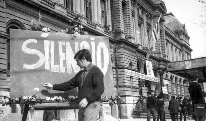 Joven en la puerta de la Facultad de Derecho frente a un cartel que dice "Silencio"