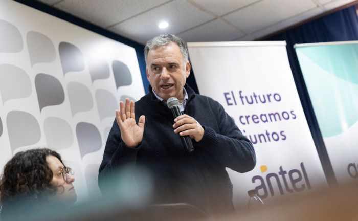 El presidente Yamandú Orsi habla, de pie, sosteniendo un micrófono con su mano izquierda, mostrando la palma de su mano derecha. Detrás, un banner con el logo de Antel y la leyenda "El futuro lo creamos juntos"