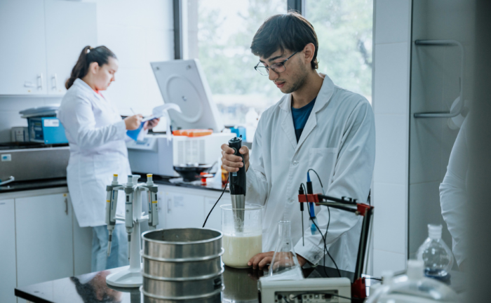 Estudiante de UTEC, en laboratorio, manipula instrumento en recipiente con leche. Detrás, una estudiante escribe en un cuaderno