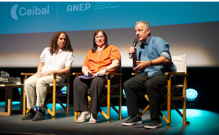 Director nacional de Educación, habla sentado con un micrófono de mano. A su lado, la presidenta de Ceibal, Fiorella Haim, y la consejera de ANEP, Elbia Pereira