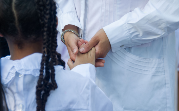 Niña de trenzas, con túnica, toma la mano de una maestra