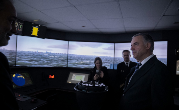 Armada Nacional: Presidente Orsi participó en reconocimiento a contralmirantes y visitó centro de simulación Presidente Yamandú Orsi de pie en el centro de simulación de la Escuela Naval, escucha atentamente a funcionario de la Armada Nacional. De fondo, las pantallas del simulador reproducen un mar