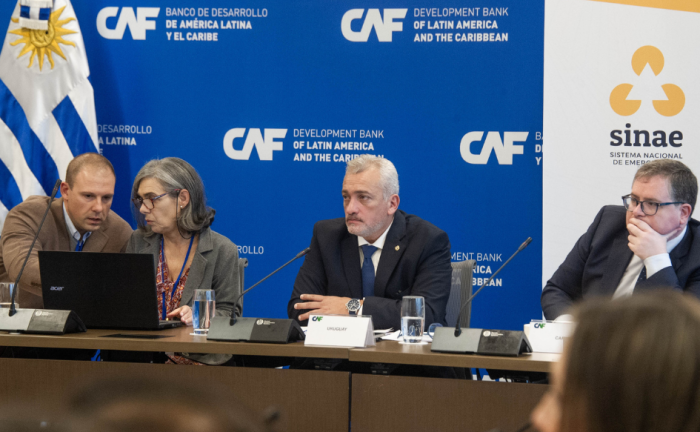 Director del Sinae, Leandro Palomeque, preside la reunión del grupo de trabajo sobre desastres naturales del Consenso de Brasilia. A su lado, dos personas hablan mirando una notebook. Una de ellas, la señala. Detrás, el pabellón nacional, un back con el logo de CAF y un banner con el logo del Sinae