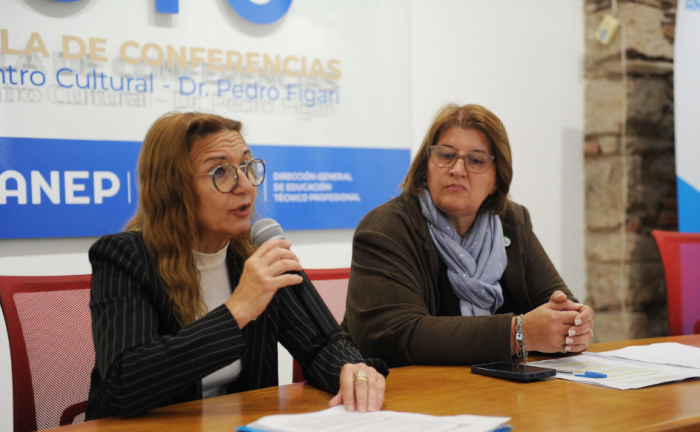 Directora del INR, Ana Juanche, habla con un micrófono en la mano. A su lado, la observa la directora de UTU, Virginia Verderese