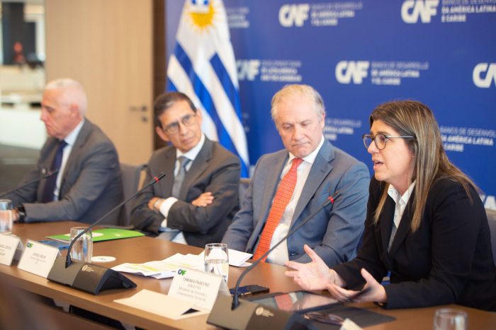 Ministra Paseyro diserta sentada, con sus brazos apoyados en una mesa. A su lado, autoridades de CAF y el ministro del Interior, Carlos Negro, la miran atentamente. De fondo, el pabellón nacional.